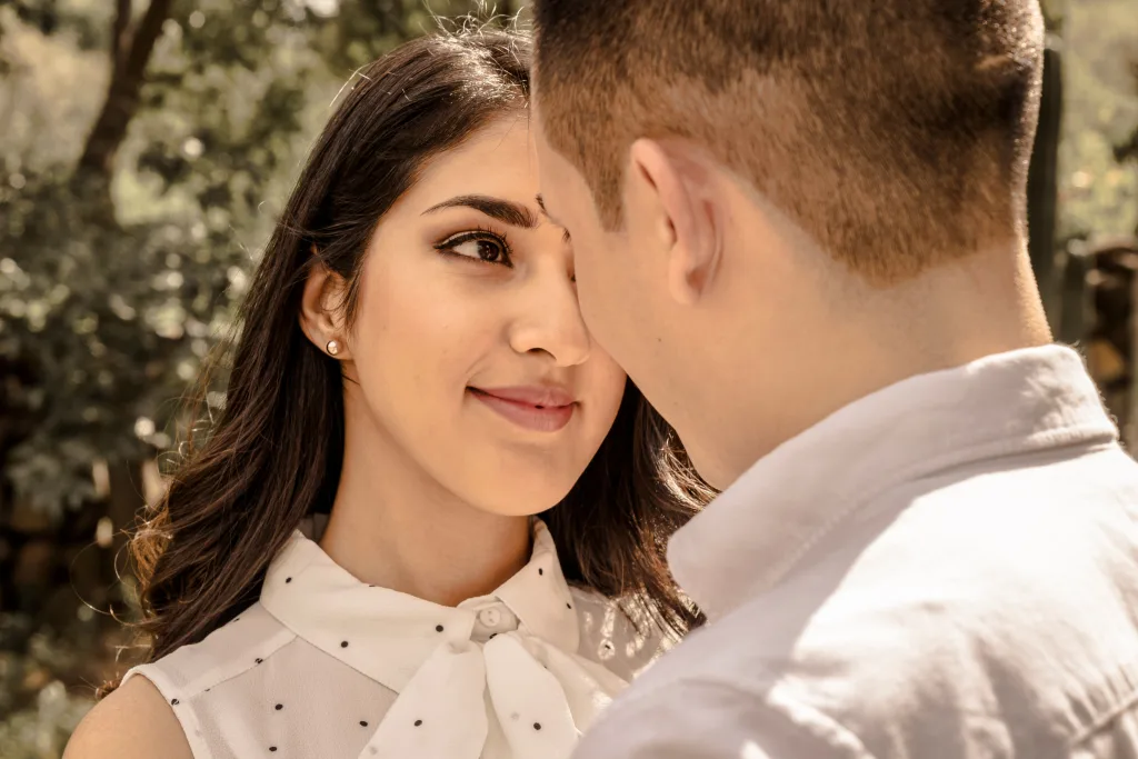 A young couple stands face to face, sharing a loving moment under a tree in daylight