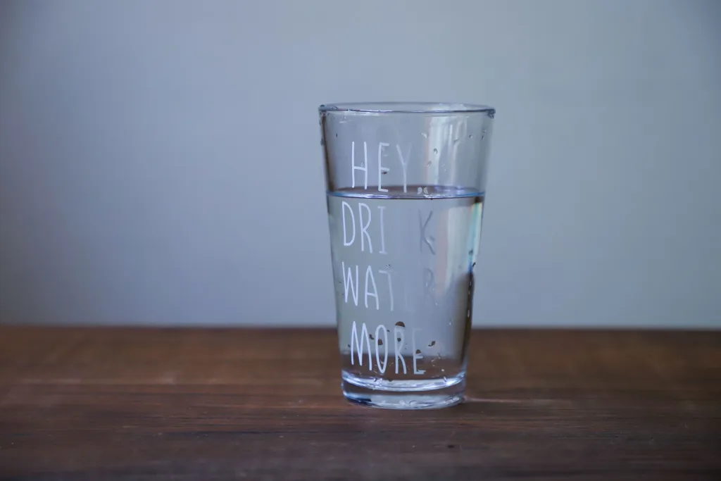 A glass of clear water on a wooden table with the text 'HEY DRINK WATER MORE'